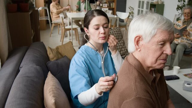 Female nurse in blue scrubs using stethoscope to examine lungs of senior man in retirement home. Footage suits geriatric healthcare promotions and medical ads