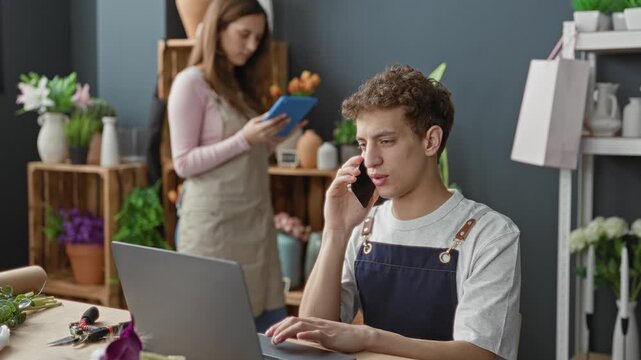 Man wearing apron holds smartphone to ear while typing on laptop in flower shop building; teamwork focus.