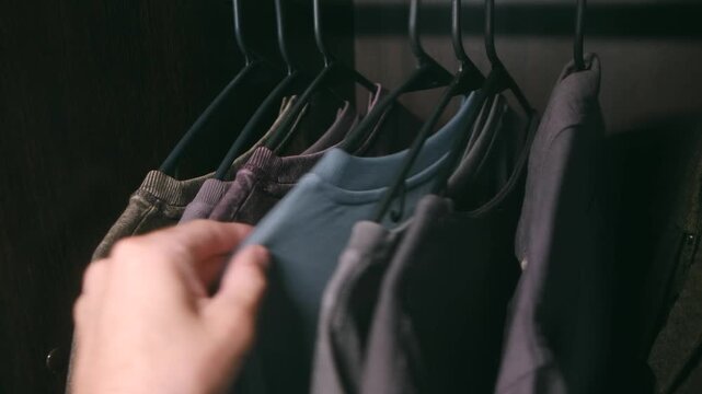 Man hand browsing through row of casual t shirts and sweatshirts hanging on rack in dark closet, searching for right outfit for day before making final selection. Person choosing clothes from wardrobe