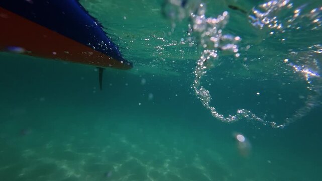 The orange paddle blade enters the turquoise water, creating bubbles, splashes, and shimmering reflections beneath the surface. A cinematic perspective of stand up paddleboarding on a sunny summer day