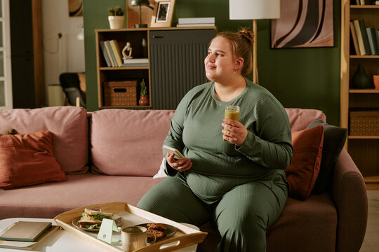 Young plus size Caucasian woman sitting on sofa, holding phone and smoothie while snacking at home, representing mindful routine and self care. Ideal for wellness marketing and lifestyle campaigns