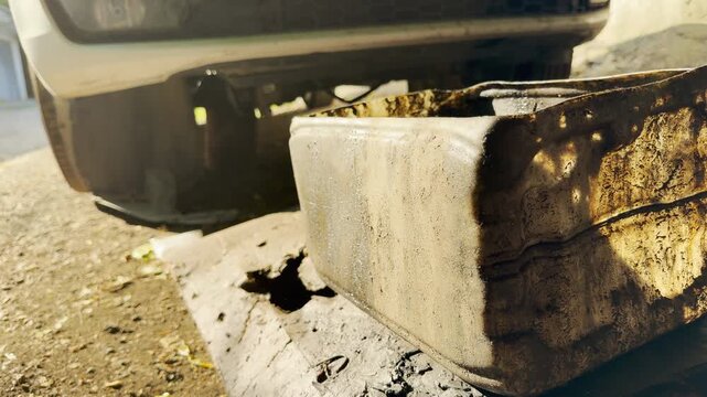 A messy plastic canister catches dirty used engine oil. The greasy container sits under a white car bumper, giving a weary sense of hard work.