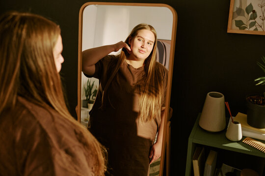 Young plus size Caucasian woman adjusting hair while viewing reflection in mirror, expressing body confidence and self acceptance. Usable for beauty, wellness, self esteem campaigns