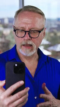 Bearded Caucasian male in glasses uses smartphone for video chat. Man in blue shirt talks to someone via the internet. Close up. Vertical video