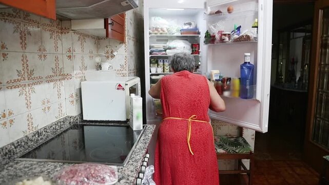  Mujer mayor buscando ingredientes dentro de la cocina del refrigerador de su casa. Abuela mayor buscando ingredientes en el refrigerador de la cocina de su casa. Aut&eacute;ntica rutina diaria de cocina 