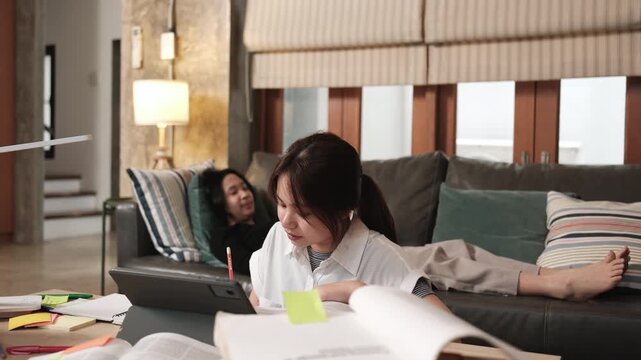 Two Asian teenage tutorials, girl concentrates on online class, remote learning at home with tablet, her friend uses mobile phone on sofa, self-study with textbooks, homeschooling together lifestyle.