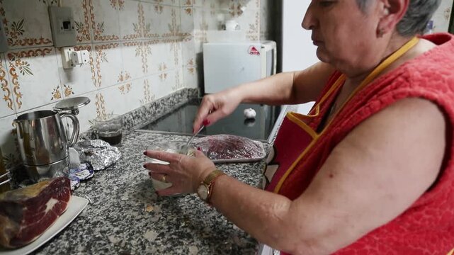  Mujer mayor remojando pan en un recipiente de vidrio lleno de leche. Abuela mayor remojando pan en un recipiente de vidrio con leche. Preparaci&oacute;n de recetas tradicionales de alb&oacute;ndigas