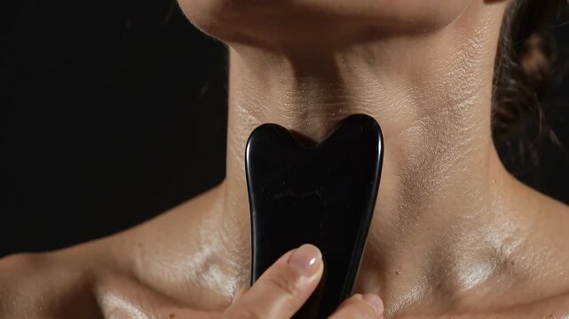 Extreme close-up of a woman massaging her neck and collarbone with a black jade gua sha stone tool for lymphatic drainage.