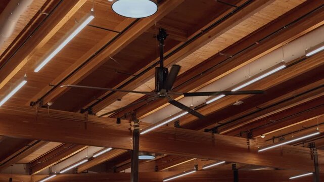Static upward view of a large ceiling fan slowly rotating beneath exposed wooden beams indoors.