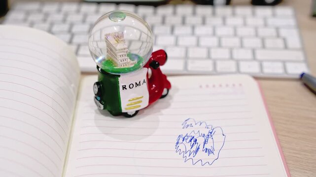 Close-up of a small Rome scooter souvenir snow globe resting on an open lined notebook with blue ink scribbles. A blurred white computer keyboard sits in the background on a modern office desk.
