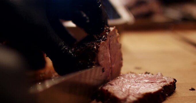 Close up shot of hands in black gloves slicing roasted meat on wooden cutting board