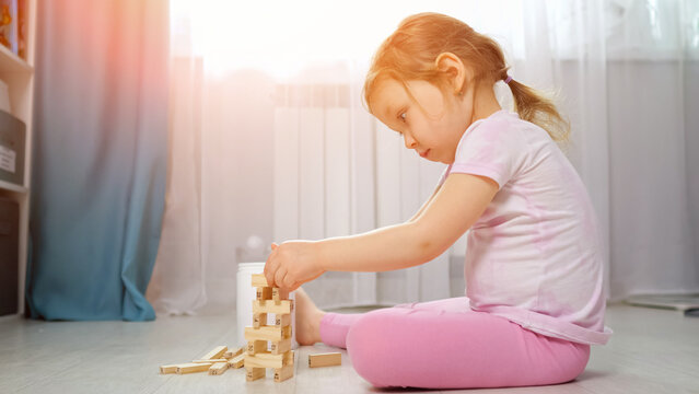 Toddler girl sitting on the floor indoors, intently concentrating on stacking and balancing wooden building blocks, engaging in childhood play for learning and development