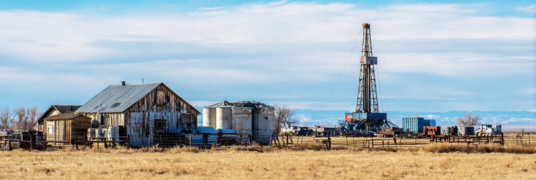 A drilling rig stands tall in a rural area with surrounding outbuildings and tanks. The sky is clear, showing a typical workday in oil or natural gas extraction