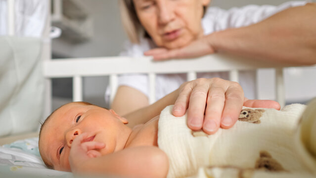 Grandmother's hand gently touching the swaddled newborn baby as it rests in a crib, showing generations connecting with care, love, and protection in a home environment