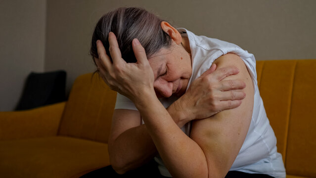 Woman sitting on a sofa, covering her face and holding her arm, expressing emotional pain, sorrow, and being a victim of domestic violence and abuse, highlighting trauma and fear at home
