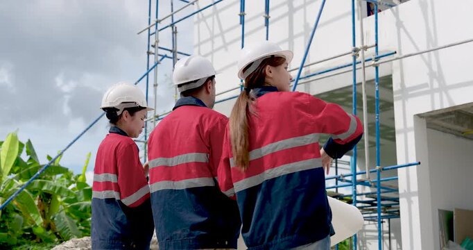 Asian woman engineer points toward scaffold and upper facade while male contractor watches, leading construction safety compliance discussion, building progress review, site supervision meeting