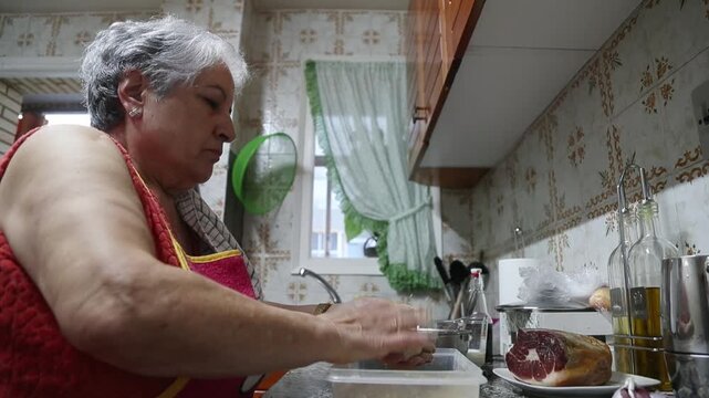  Mujer mayor rebozando alb&oacute;ndigas caseras en un bol de pan rallado. Abuela mayor enrollando y rebozando alb&oacute;ndigas en pan rallado. Receta casera tradicional, aut&eacute;ntica cocina senior.
