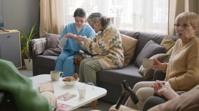 Female caregiver helping senior woman with knitting in nursing home while other seniors socialize. Video suitable for senior care services and retirement community marketing