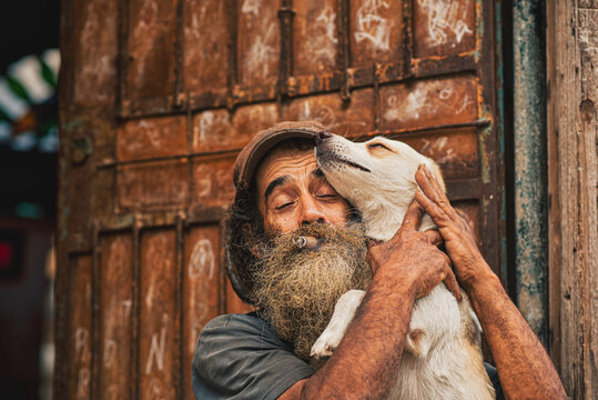 Bearded Cuban man holding his beloved dog while smoking a traditional cigar in Old Havana, Cuba. The portrait captures the bond between a pet owner and his companion animal