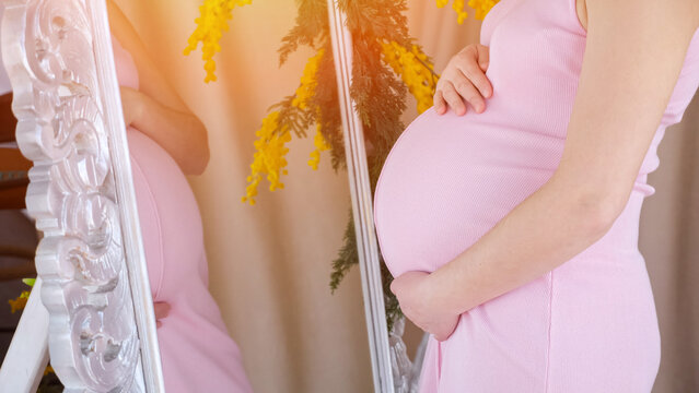 Pregnant woman in a pink dress gently touching her baby bump, observing her reflection in an ornate mirror, symbolizing motherhood, anticipation, and the bond with her unborn child