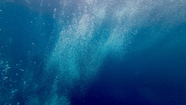 Air bubbles rise from the abyss through deep blue water. Abstract underwater background with shimmering oxygen stream and dramatic natural texture.