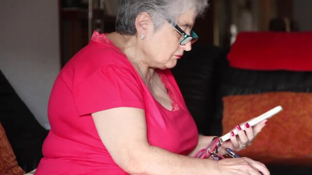  Mujer mayor con bata rosa usando un tel&eacute;fono inteligente en el sof&aacute; de su casa. Mujer mayor con gafas navegando por el tel&eacute;fono inteligente en casa. Senior activo, estilo de vida tecnol&oacute;gico moderno.