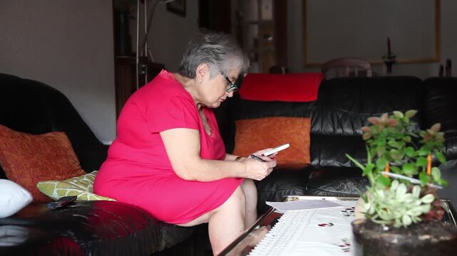  Mujer mayor con bata rosa usando un tel&eacute;fono inteligente en el sof&aacute; de su casa. Mujer mayor con gafas navegando por el tel&eacute;fono inteligente en casa. Senior activo, estilo de vida tecnol&oacute;gico moderno.