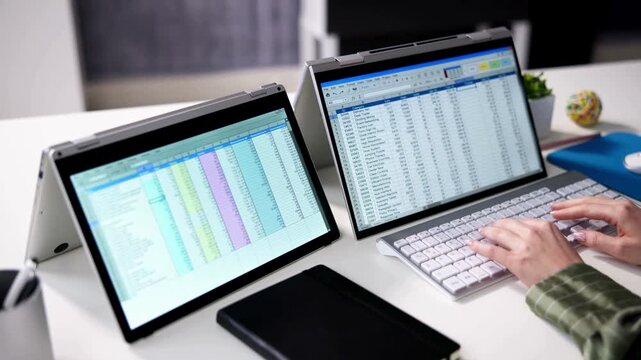 Professional data analyst working with spreadsheets on dual laptop screens, financial accounting software on multiple monitors, corporate office workspace