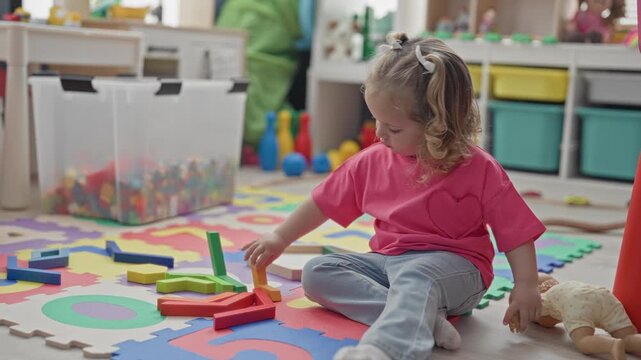 Girl reaches with hand to stack colorful wooden blocks and holds a doll on a foam mat in a playroom inside a building; curiosity calm.