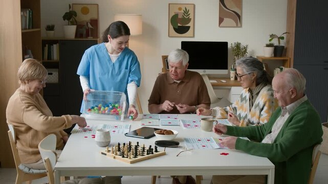 Young female caregiver in blue scrubs hosting bingo game for diverse group of seniors in retirement home. Video for promoting geriatric healthcare and assisted living services