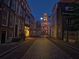 Dordrecht, South Holland, Holland A lively European square at dusk, featuring historic buildings with glowing windows, street lamps, and outdoor cafes. People sit on benches. © Sami