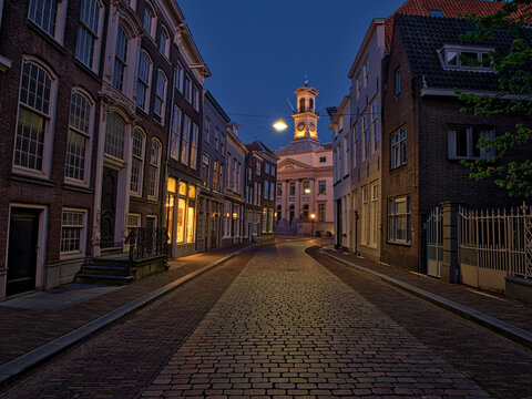 Dordrecht, South Holland, Holland A lively European square at dusk, featuring historic buildings with glowing windows, street lamps, and outdoor cafes. People sit on benches.