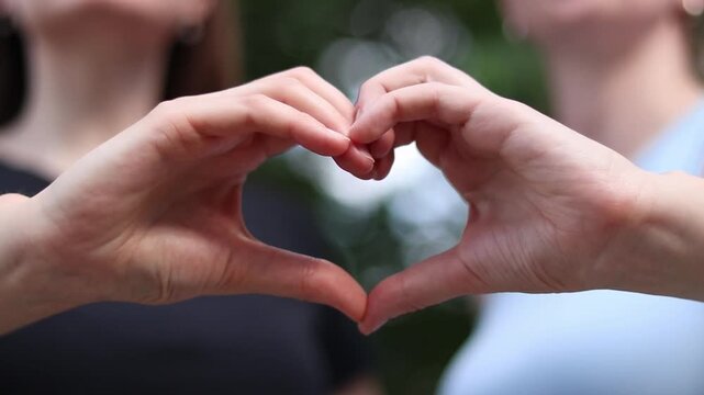 Two female hands make a heart gesture on a green park background. Close-up of hands forming a heart symbol with fingers on a blurred natural background. Concept of love and positive emotions