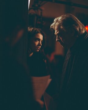 A young actress and an older actor exchange a tense look on a dimly lit film set, with soft key light, shallow depth of field, script pages, and a camera rig creating a dramatic behind-the-scenes