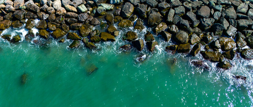 Top down aerial drone perspective of ocean water meeting the rugged stones of a breakwater