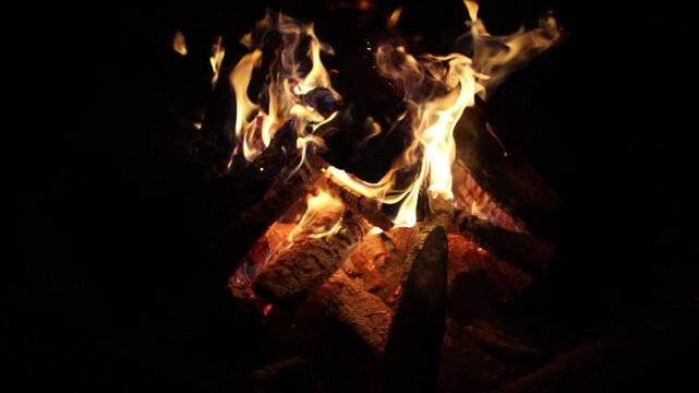 Bright flames and glowing embers burn through stacked wooden logs against a dark night background, creating a warm dramatic campfire scene with copy space