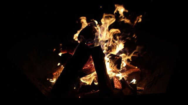 Bright flames and glowing embers burn through stacked wooden logs against a dark night background, creating a warm dramatic campfire scene with copy space
