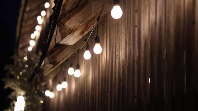 Glowing string light bulbs suspended against a rustic wooden slat wall at night, creating a warm, cozy ambiance with shallow depth of field and soft bokeh