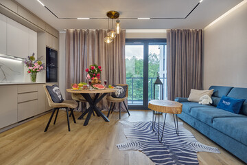 Salon w nowoczesnym apartamencie z beżowym  aneksem kuchennym z drewnianym stołem zastawionym kwiatami i śniadaniem © arteffect.pl