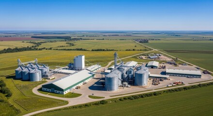 Fototapeta premium Modern agricultural processing facility with grain silos and storage buildings in a vast rural landscape under a clear blue sky.