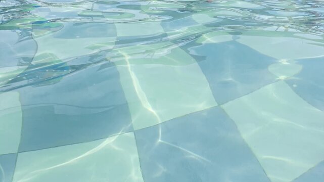 clear water rippling gently in a swimming pool. The sunlight creates shimmering, dancing light patterns (caustics) on the bottom, which is lined with light blue and teal checkered tiles.