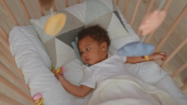 African american girl reaches with hand for plush mobile above wooden crib in nursery while holding a teether and blanket; serenity.