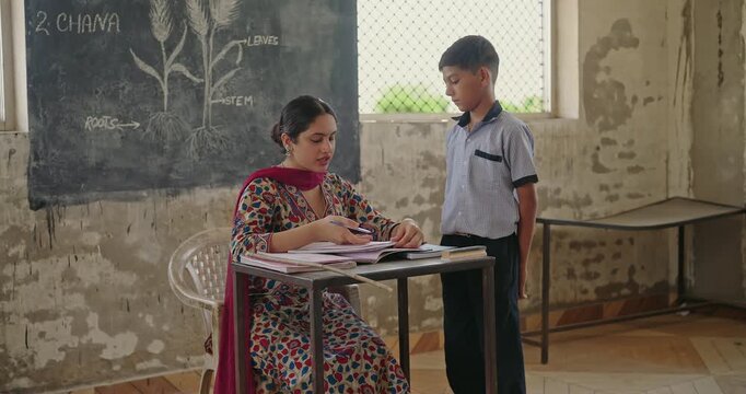 Indian young adult female sitting on chair do work check study book talk speak at class room rural area place. Strict Tutor look angry hold stick hitting teen boy child hand for project notes mistake
