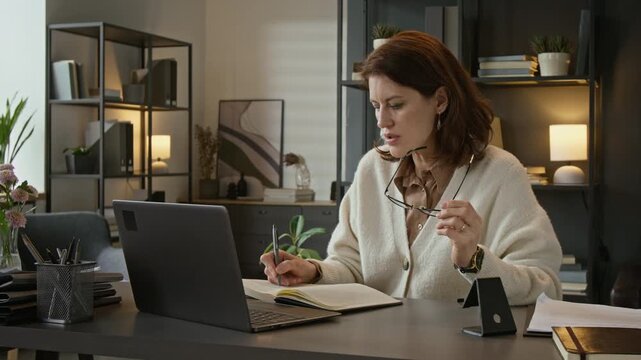Medium shot of middle aged Caucasian female psychotherapist talking during online therapy session on laptop and writing notes in notebook. Suitable for mental health and telehealth advertising videos