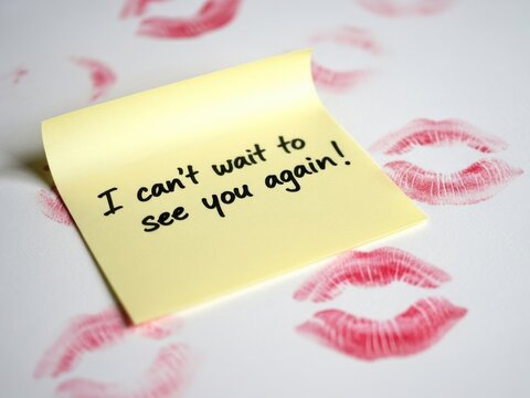 Yellow sticky note with handwritten message i cant wait to see you again on a background of red lipstick kisses