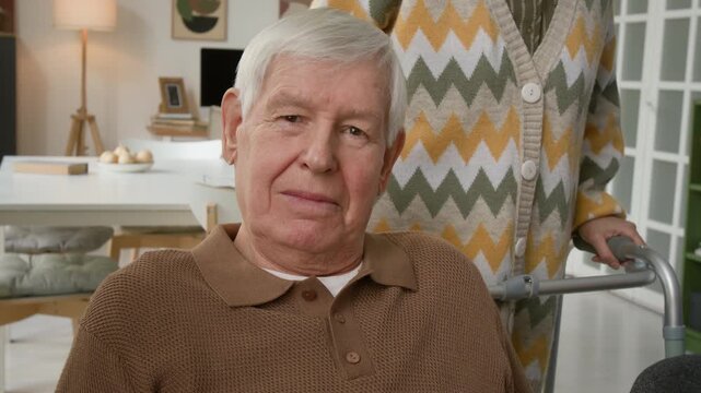 Portrait of senior man sitting in retirement home with senior woman holding walker. Footage for promoting geriatric healthcare, assisted living, and senior care services