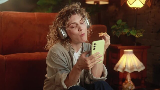 Horizontal video of curly haired woman wearing headphones, eating pizza and casually scrolling smartphone during relaxing break at home. High quality 4k footage