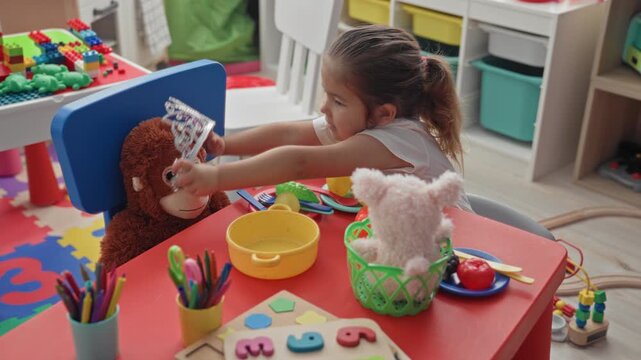 Girl places tiara on stuffed monkey at play table in building playroom, hands reaching toward toy; joyful imaginative play.