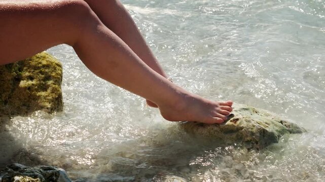 Close-up of bare female feet with a shell anklet on wet rocks as sea waves wash over them, handheld summer shot.