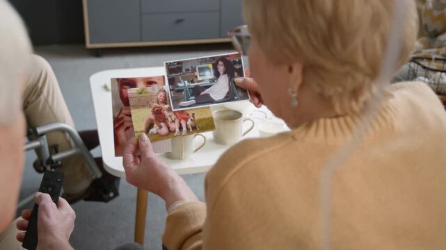 Over shoulder shot of senior woman looking at printed family photographs while sitting together in nursing home living room. Retirement community marketing or memory care